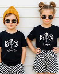 Two children wearing black t-shirts with a skeleton design and the text "BOO Harper" printed on them, along with round sunglasses and a yellow beanie.