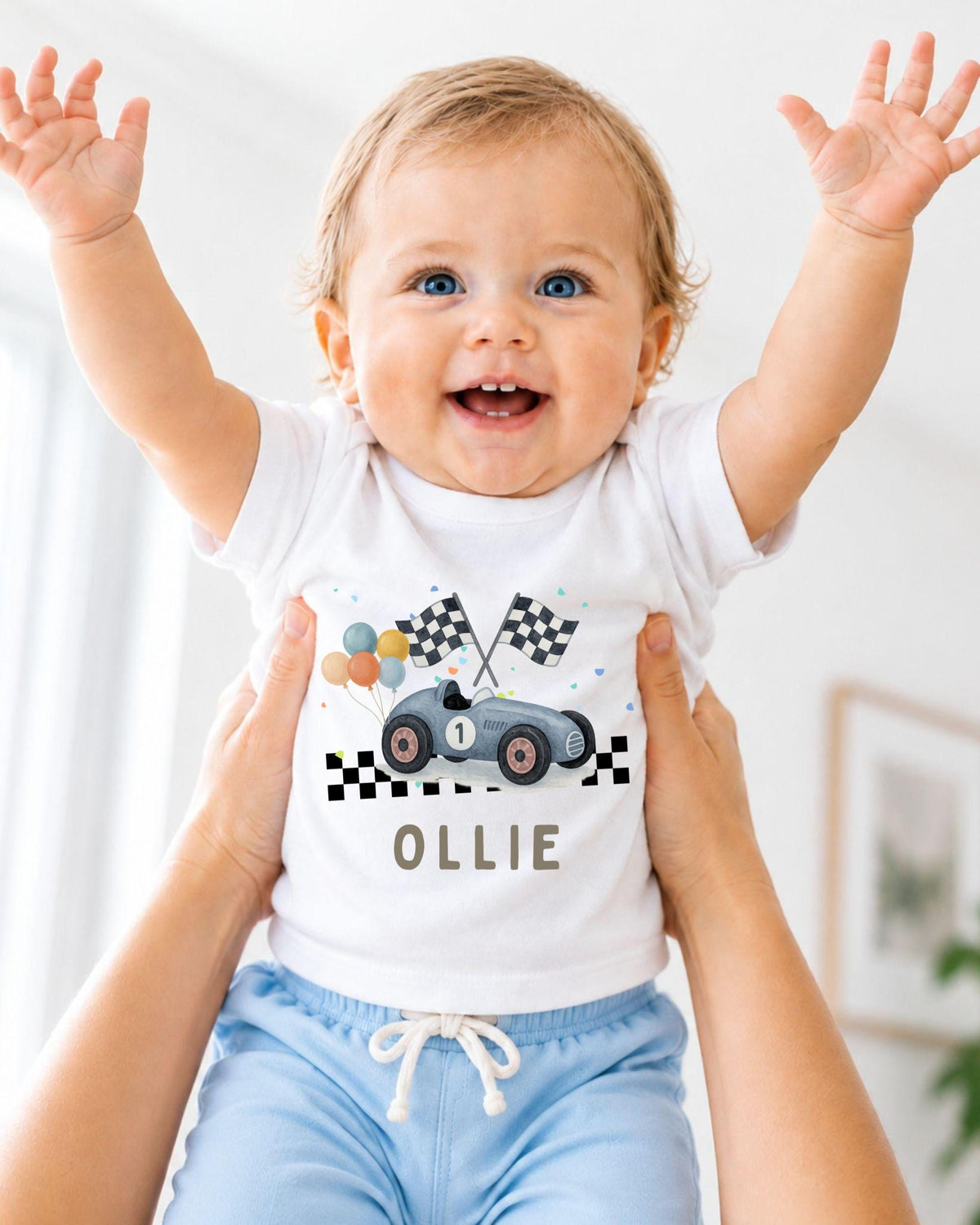 A smiling baby wearing a white t-shirt with a race car graphic and the name "Ollie" printed on it, being held up by someone's arms.