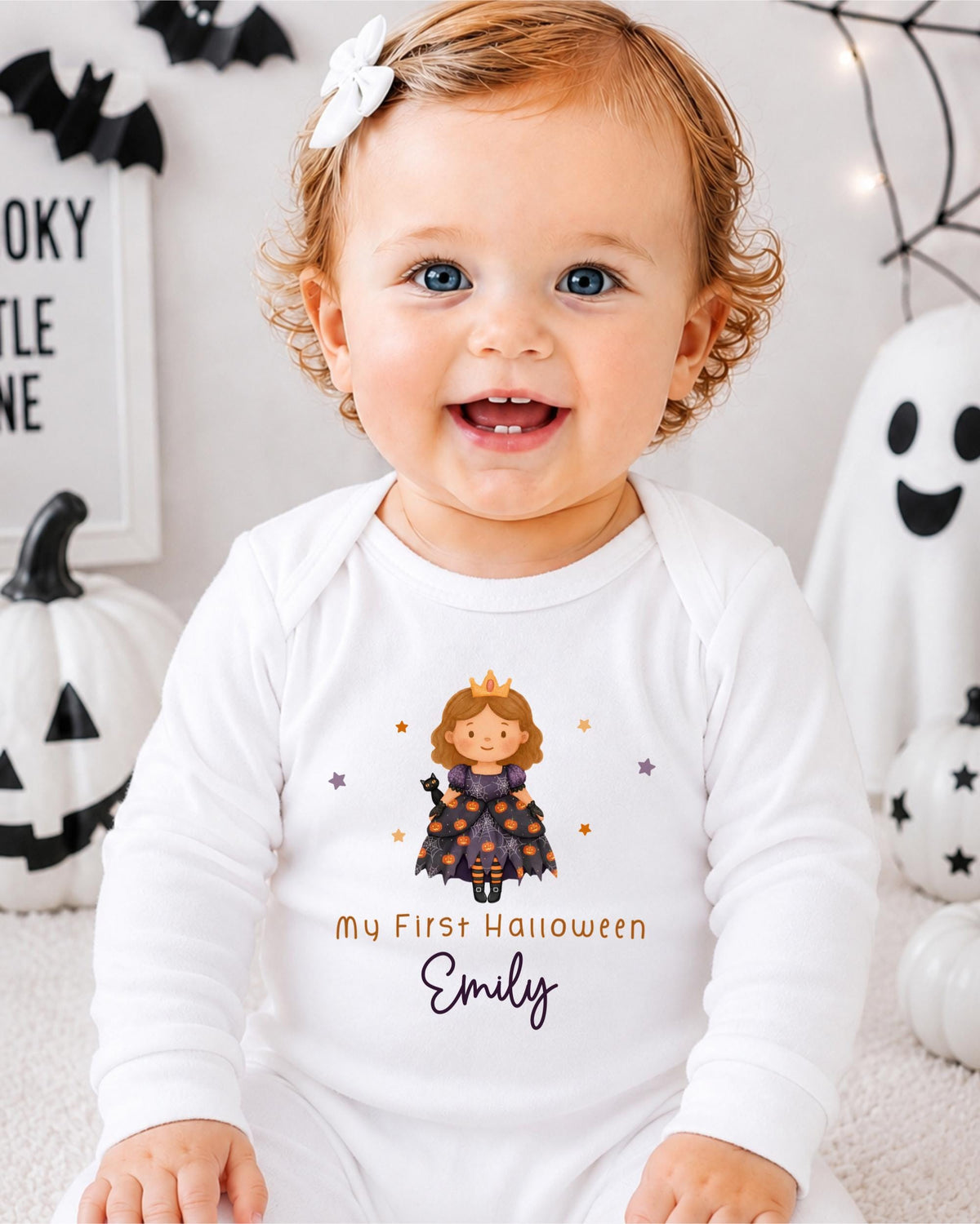 A smiling baby girl wearing a white onesie with a Halloween-themed design, sitting in front of Halloween decorations.