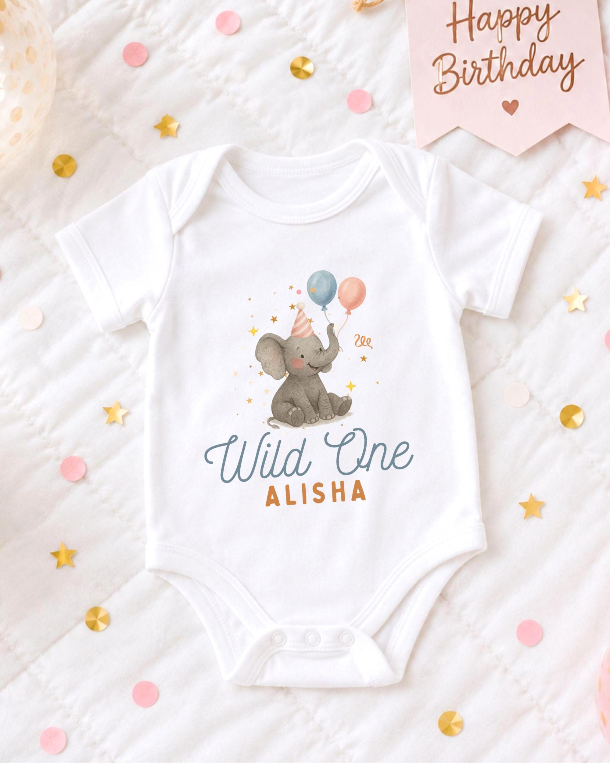A white onesie with an elephant and balloons printed on it, placed on a bed with confetti and a birthday card.