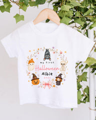 A white t-shirt with a Halloween-themed design hanging on a wooden hanger. The design includes various Halloween-related characters such as bats, skulls, and pumpkins, surrounded by a wreath of flowers and leaves. The text on the t-shirt reads "my first Halloween Albie 2020".