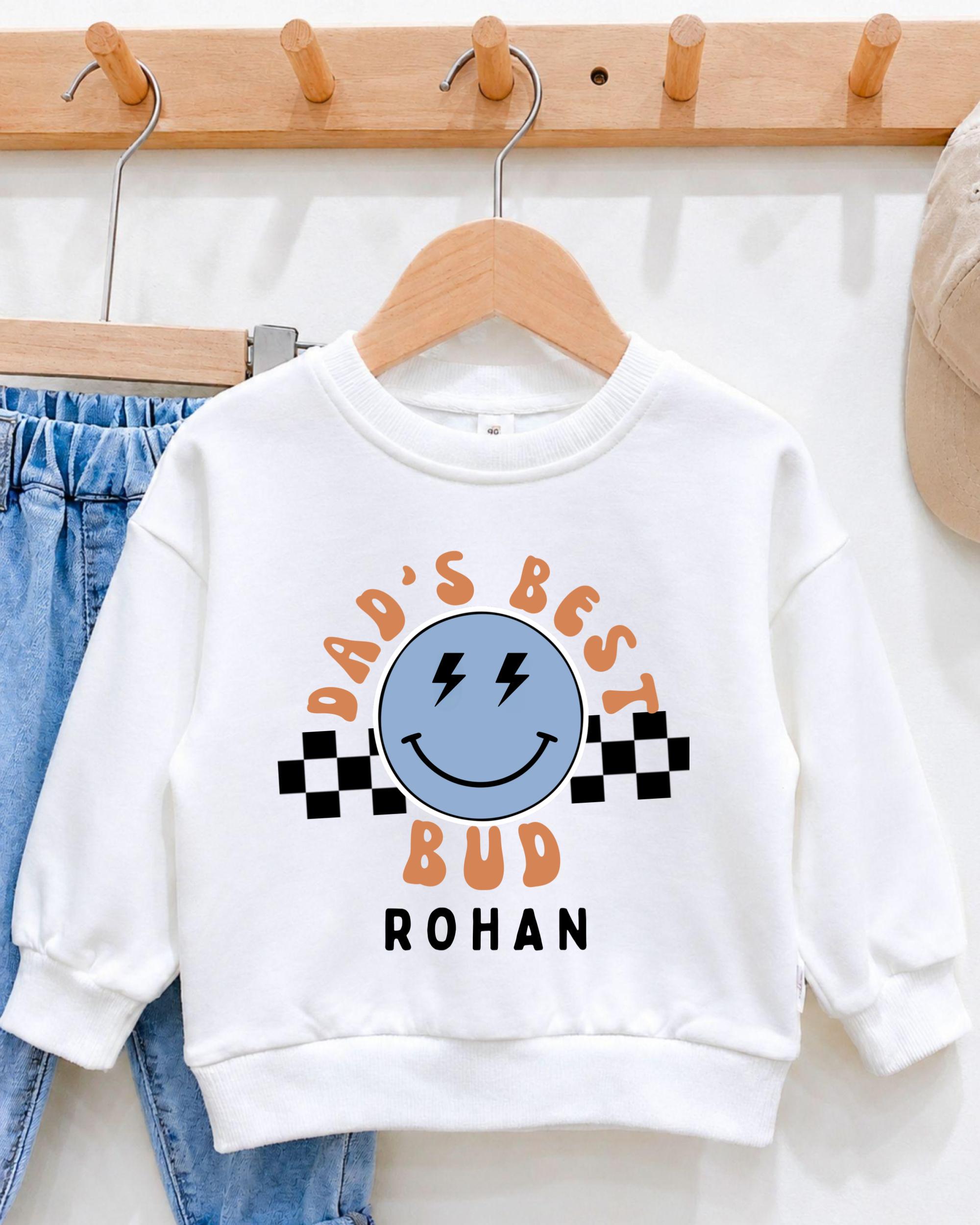 A white sweatshirt with a graphic design featuring a smiling face and text that says "DAD'S BEST BUD ROHAN".