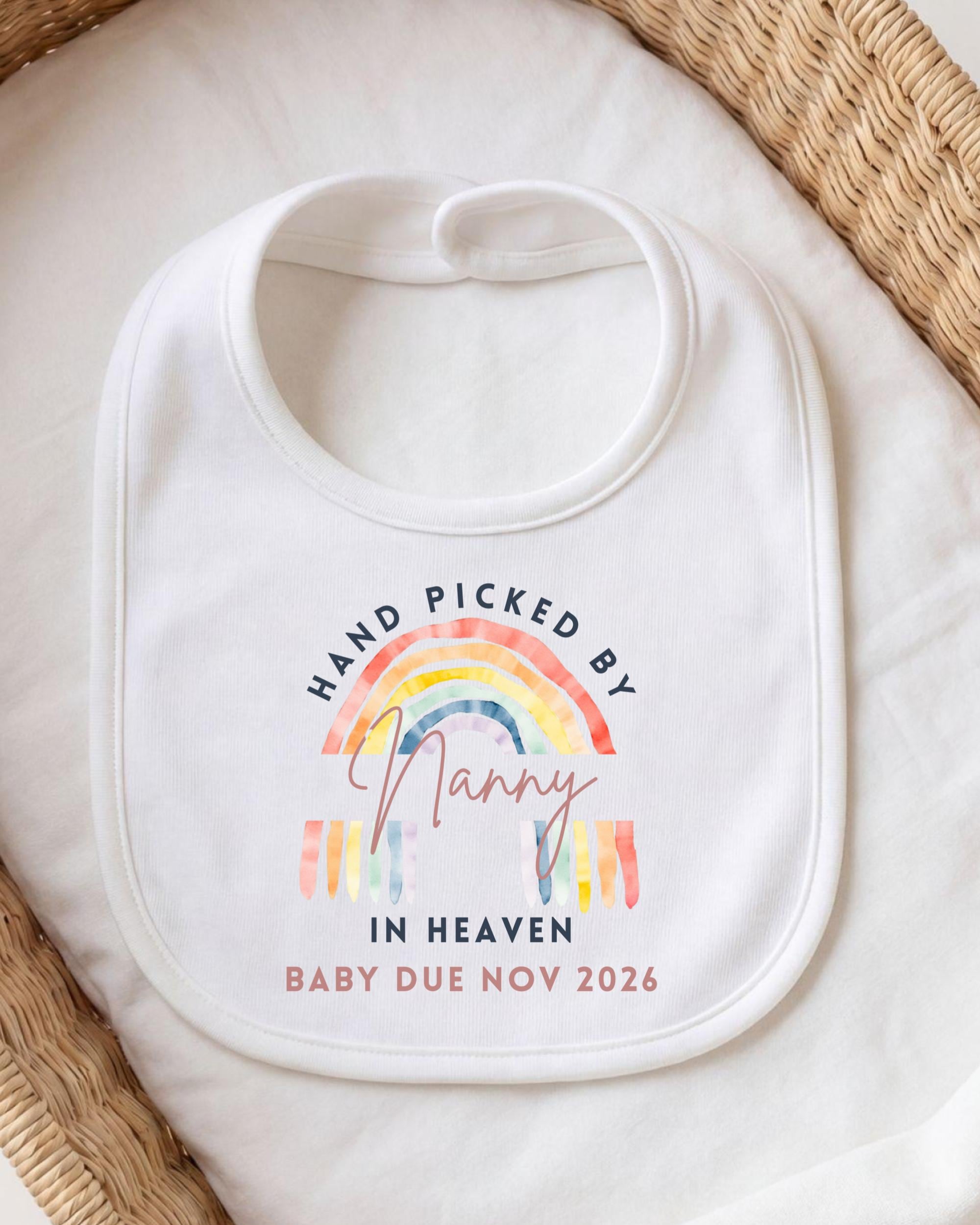A white baby bib with a colorful rainbow graphic and text that reads "Hand picked by Nanny in Heaven Baby Due Nov 2026".