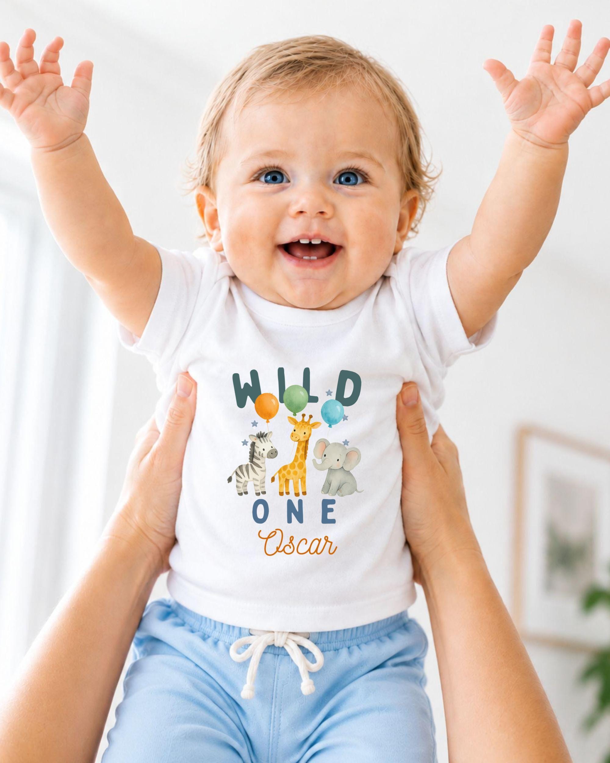 A smiling baby wearing a white t-shirt with a jungle animal design and the text "Wild One Oscar" is being held up by two people.
