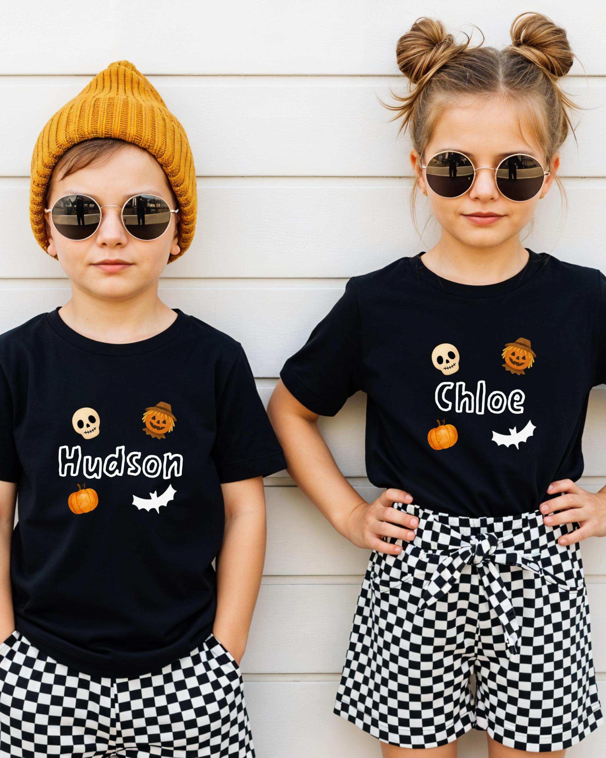 Two children wearing black t-shirts with Halloween-themed designs and sunglasses stand against a white wall.