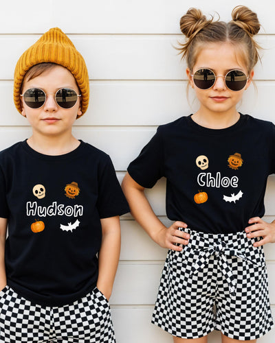 Two children wearing black t-shirts with Halloween-themed designs and sunglasses stand against a white wall.