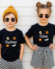 Two children wearing black t-shirts with Halloween-themed designs and sunglasses stand against a white wall.