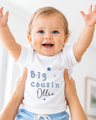 A smiling baby wearing a white shirt with the text "Big Cousin Ollie" is being held up by someone's arms.