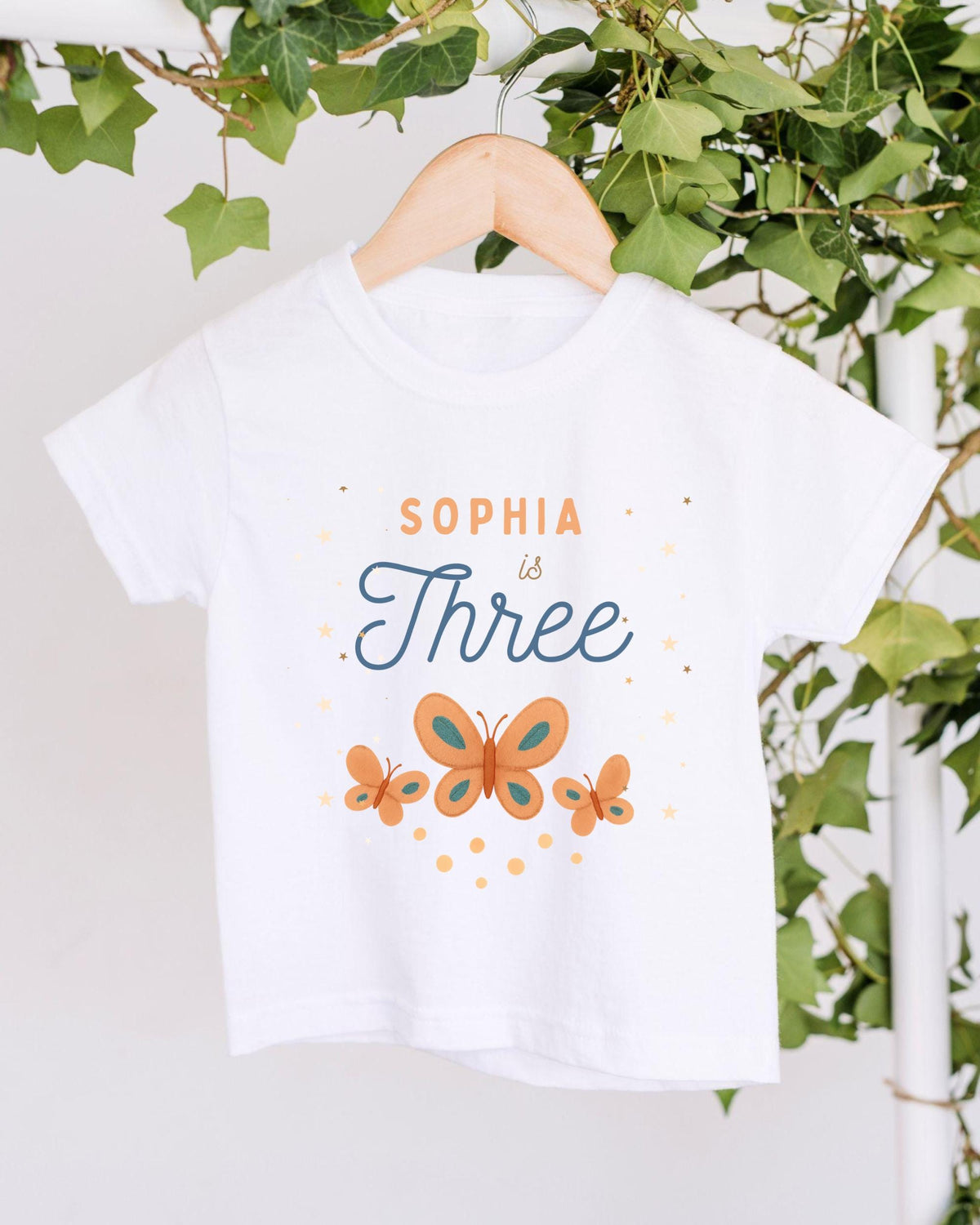 A white t-shirt with a design featuring butterflies and the text "Sophia is Three" printed on it, hanging on a wooden hanger.