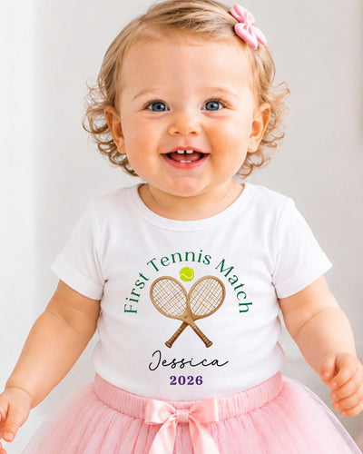 A smiling baby girl wearing a white t-shirt with a tennis racket and ball design, and a pink bow in her hair.