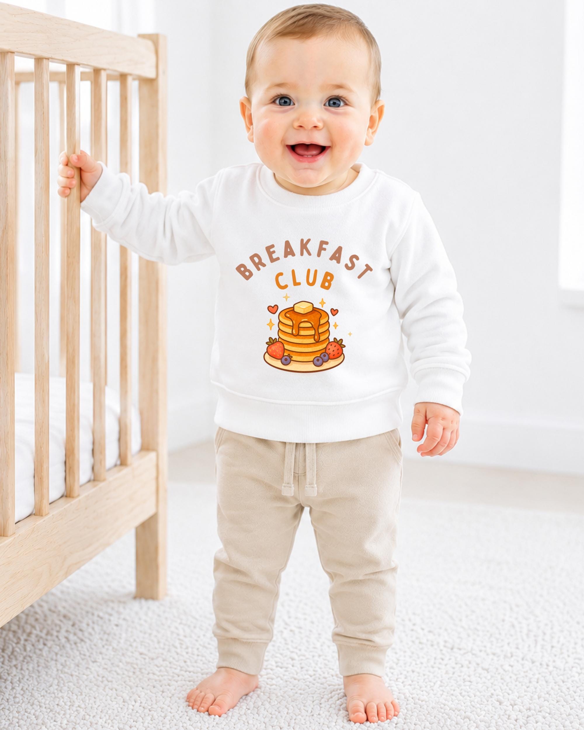A smiling baby boy wearing a white sweatshirt with the text "BREAKFAST CLUB" and an illustration of pancakes.