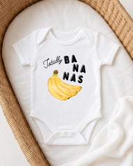 A white onesie with a banana graphic and the text "Totally BANANA" printed on it, laid out on a white blanket.
