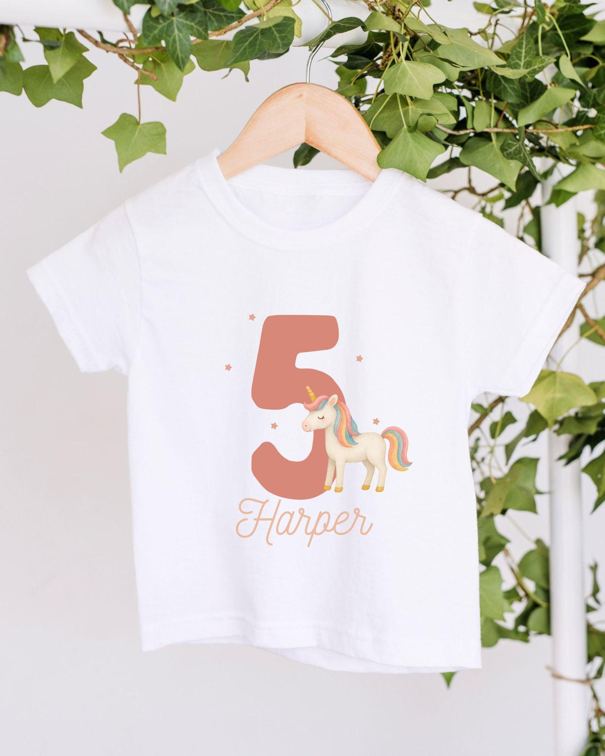A white t-shirt with a unicorn graphic and the number 5 printed on it, hanging on a wooden hanger.
