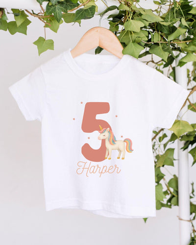 A white t-shirt with a unicorn graphic and the number 5 printed on it, hanging on a wooden hanger.