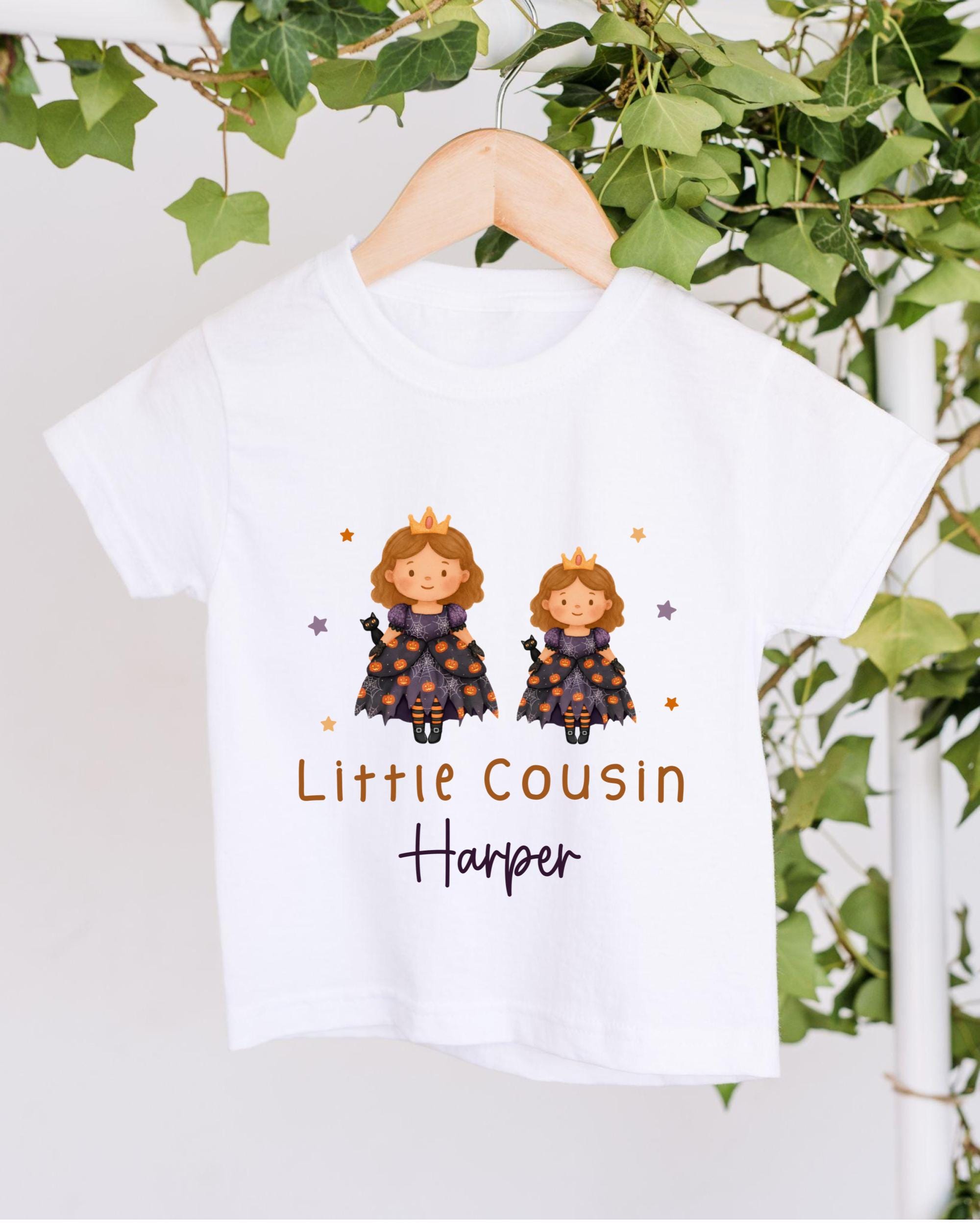 A white t-shirt with a graphic design featuring two cartoon princesses wearing crowns, surrounded by stars and vines. The text below the design reads "Little Cousin Harper".