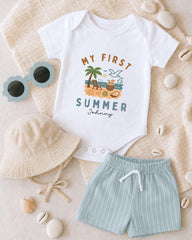 A baby's outfit, including a white onesie with a summer theme, a beige bucket hat, and light blue shorts, along with sunglasses and seashells, are arranged on a beige surface.