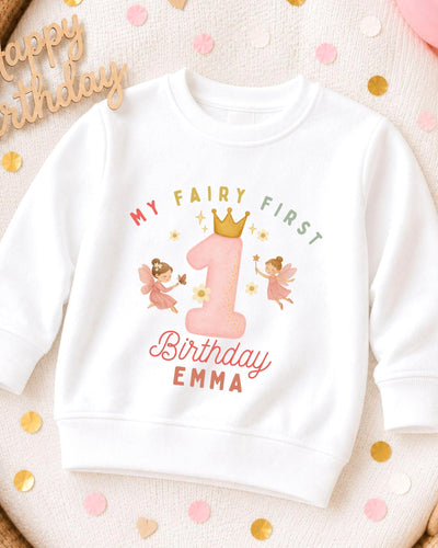 A white sweatshirt with a personalized birthday design for a child named Emma. The design features a large number one with a crown, surrounded by two pink fairy characters. The text on the sweatshirt reads "My Fairy First Birthday Emma".