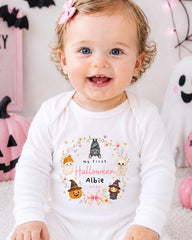 A smiling baby girl wearing a white onesie with a Halloween-themed design, sitting in front of Halloween decorations.