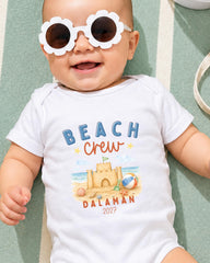 A baby wearing a white onesie with a beach-themed design and sunglasses lying on a green and white striped surface.