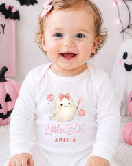 A smiling baby girl wearing a white onesie with a ghost character and the text "Little Boo Amelia" is sitting in front of Halloween decorations.