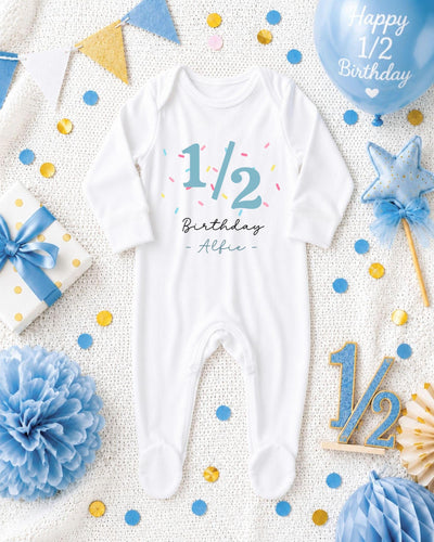 A white onesie with the text "1/2 Birthday - Alfie" printed on it, surrounded by confetti, a blue balloon, and other baby-related items.