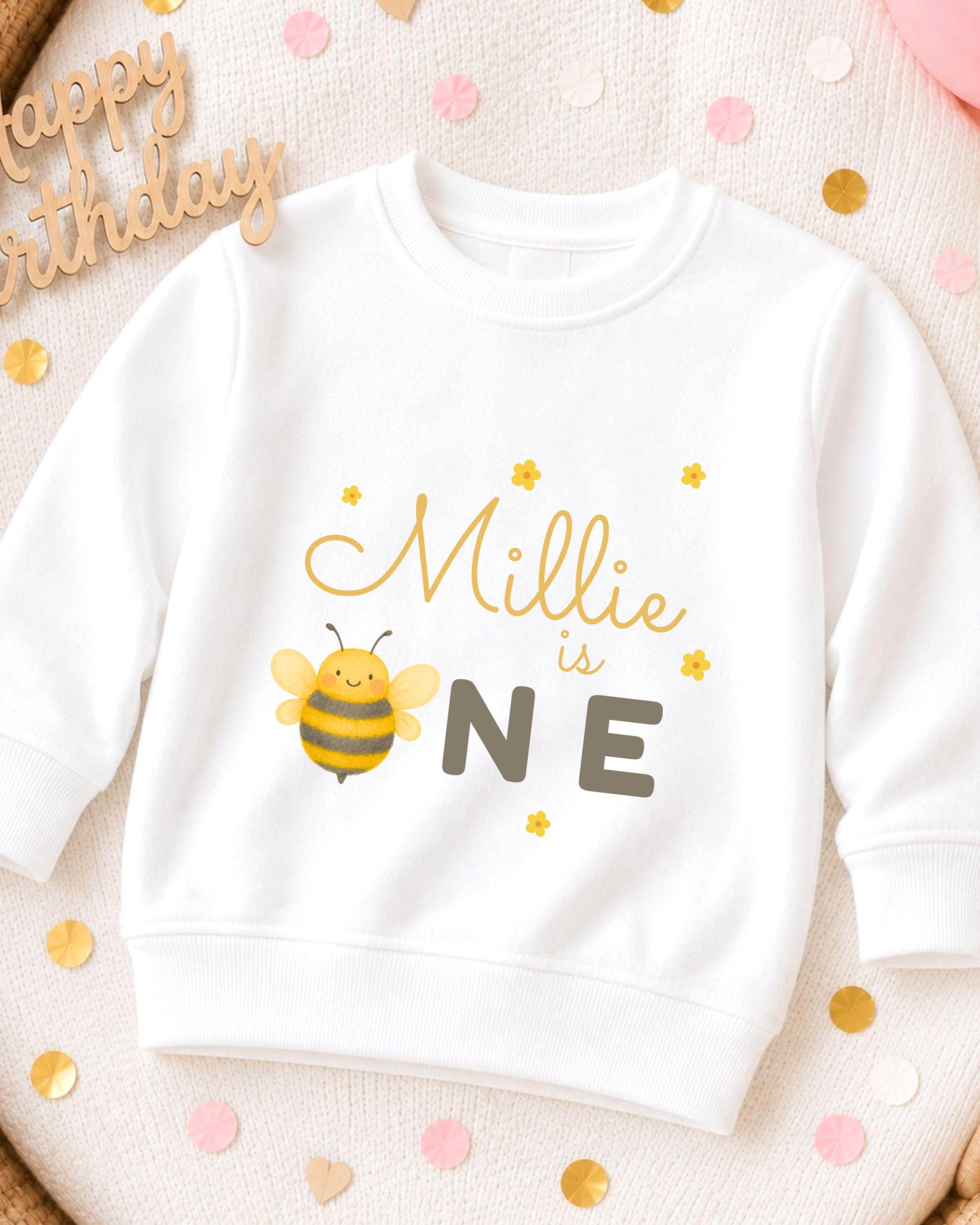 A white sweatshirt with a bee graphic and the text "Millie is one" printed on it, surrounded by confetti.