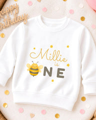 A white sweatshirt with a bee graphic and the text "Millie is one" printed on it, surrounded by confetti.