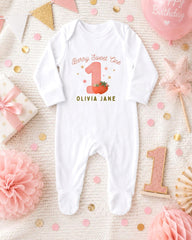 A white onesie with a pink number one and the text "Berry Sweet One Olivia Jane" printed on it, along with a pink star and confetti, is surrounded by pink and gold decorations, including a pink star, a pink flower, and a pink ribbon.