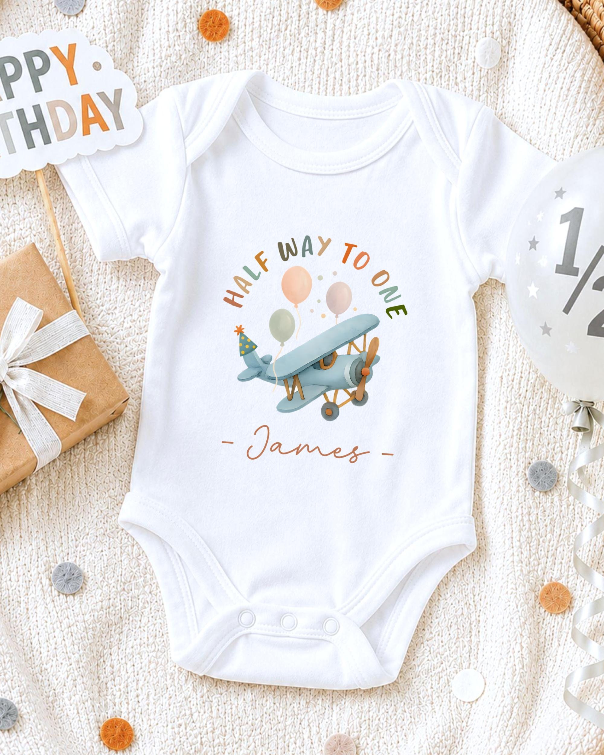 A white onesie with a cartoon airplane and the text "Half way to one - James" printed on it, along with a birthday sign and confetti.