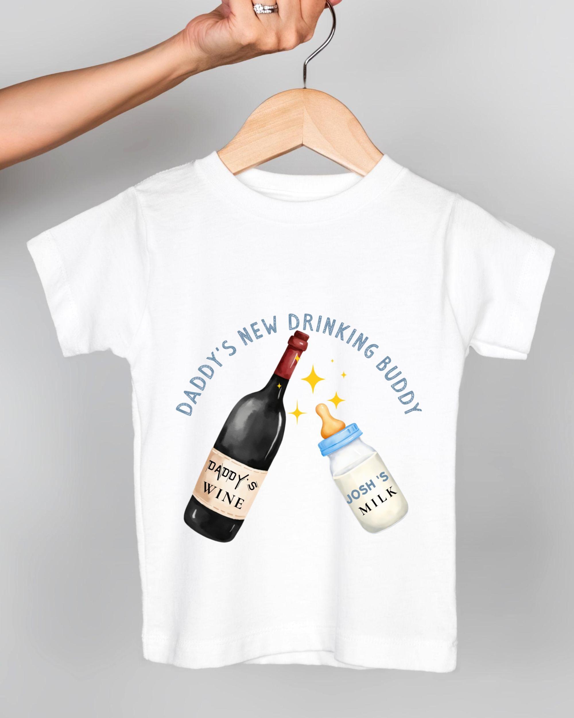 A white t-shirt with a graphic design depicting a wine bottle and a baby bottle, along with the text "DADDY'S NEW DRINKING BUDDY" and "JOSH'S MILK".