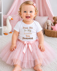 A smiling baby girl wearing a white t-shirt with text and a pink tutu sits on a white carpet surrounded by stuffed animals and a pink canopy.