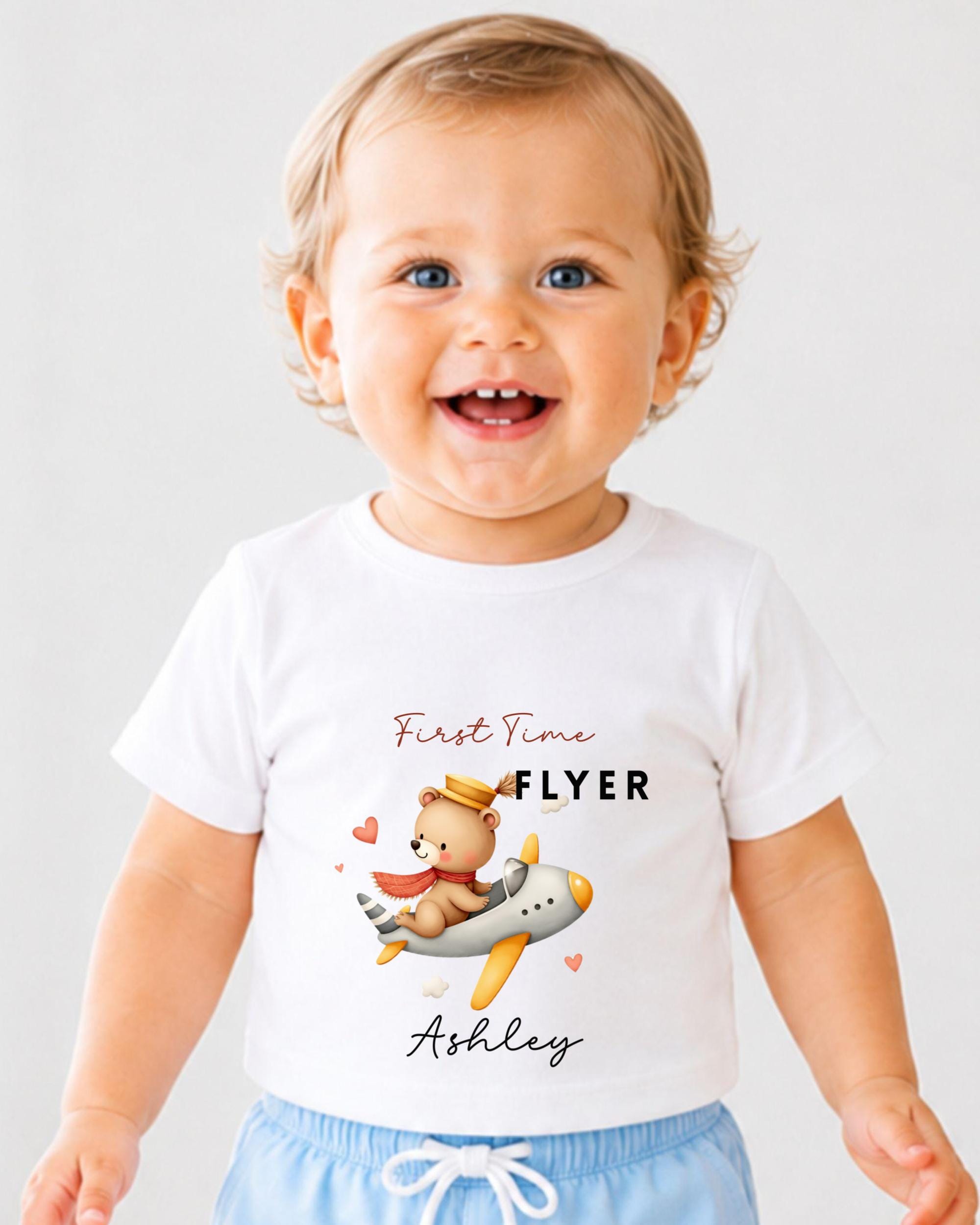 A smiling baby wearing a white t-shirt with a cartoon image of a teddy bear and the text "First Time Flyer Ashley" printed on it.
