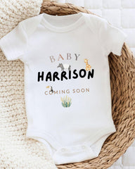A white baby onesie with the text "BABY HARRISON COMING SOON" printed on it, along with illustrations of animals.