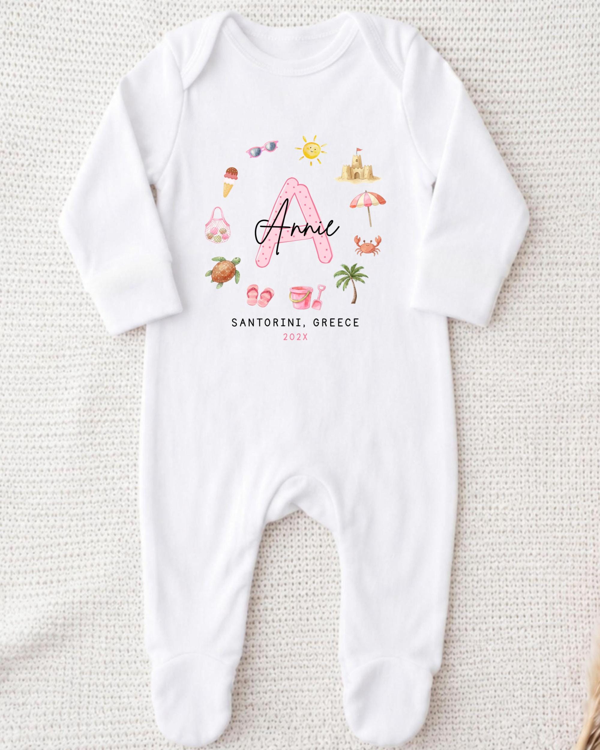 A white onesie with a personalized design featuring a pink letter "A" surrounded by various summer-themed illustrations, such as a beach, sunglasses, and a palm tree.