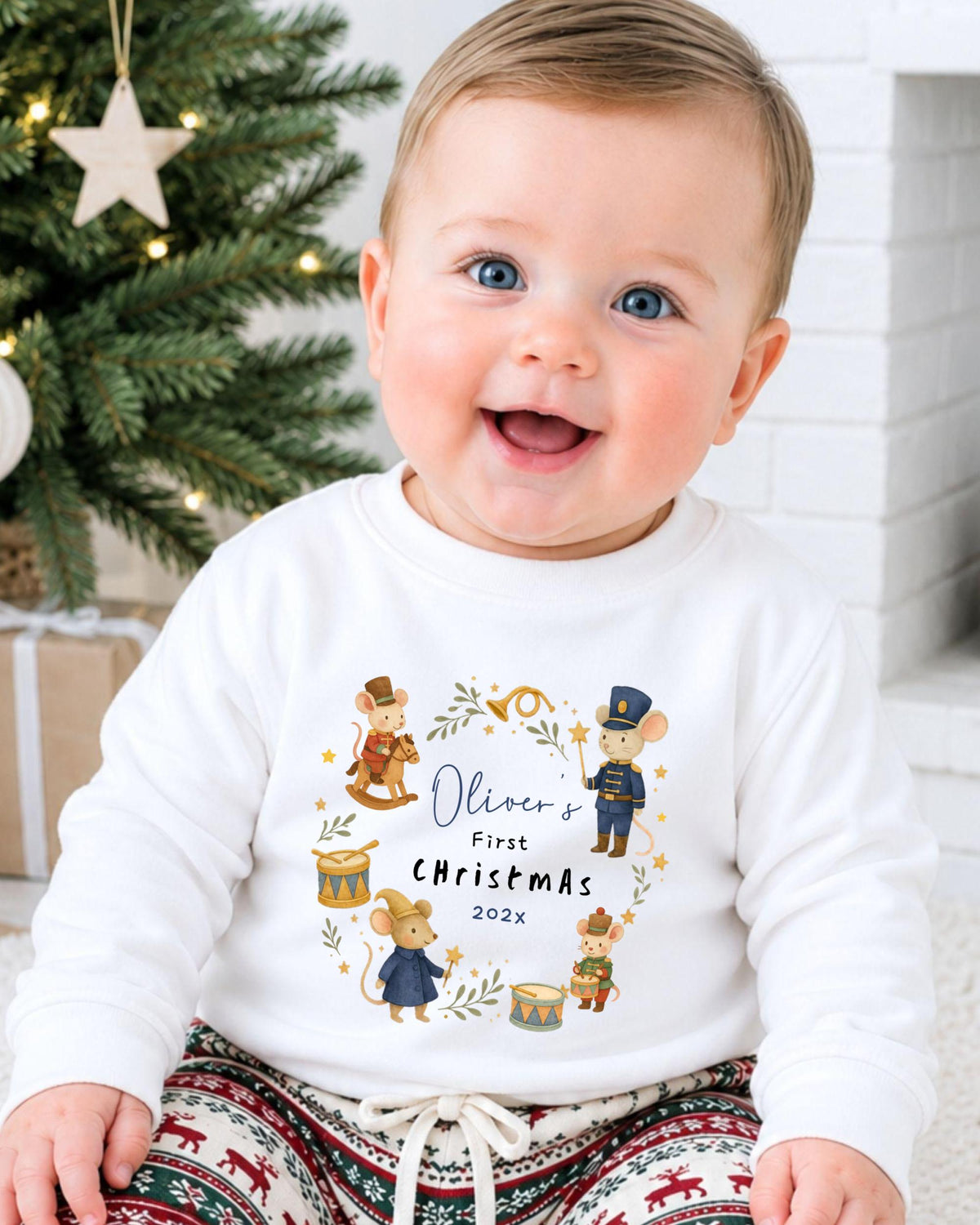 A smiling baby wearing a white shirt with a Christmas-themed design and text.