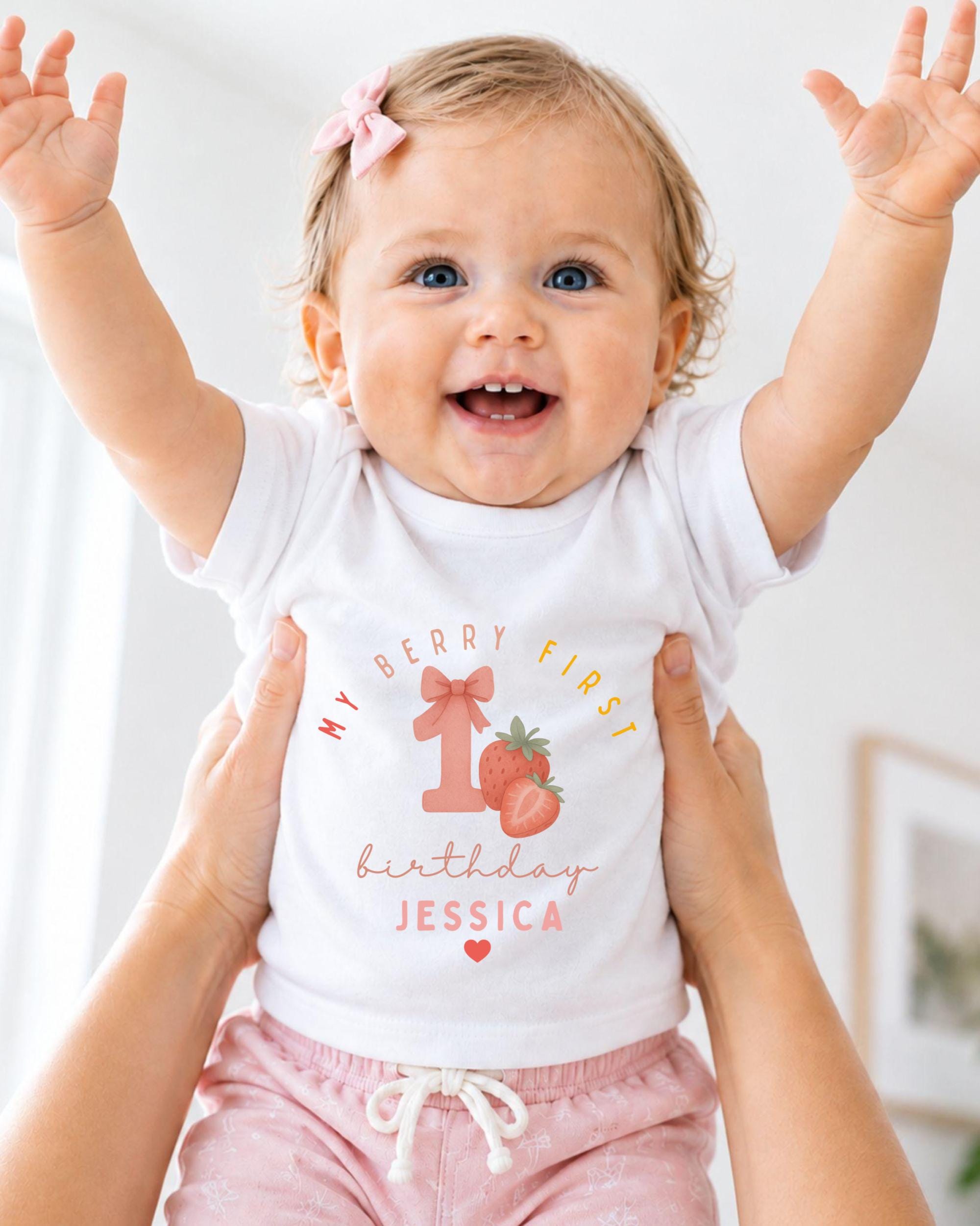 A smiling baby girl wearing a white t-shirt with a personalized design and text, being held up by someone's arms.