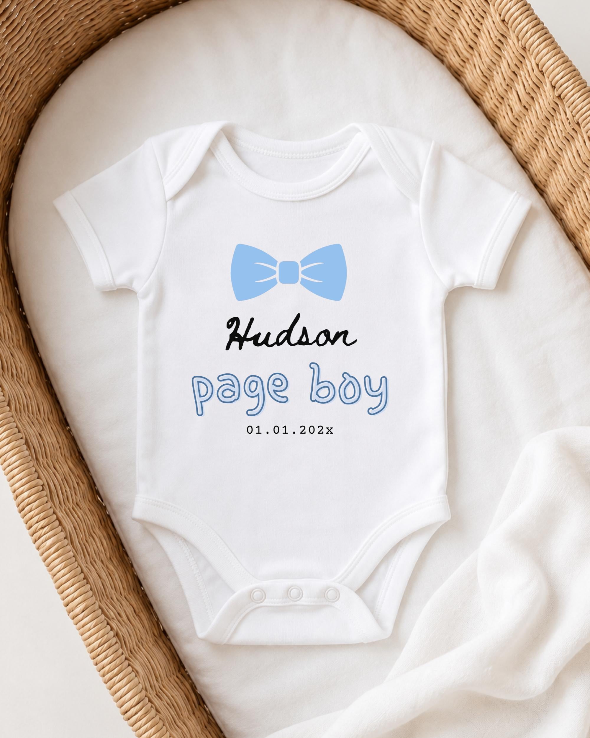 A white baby onesie with a blue bow tie and the text "Hudson page boy" printed on it, placed in a woven basket.