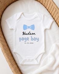 A white baby onesie with a blue bow tie and the text "Hudson page boy" printed on it, placed in a woven basket.