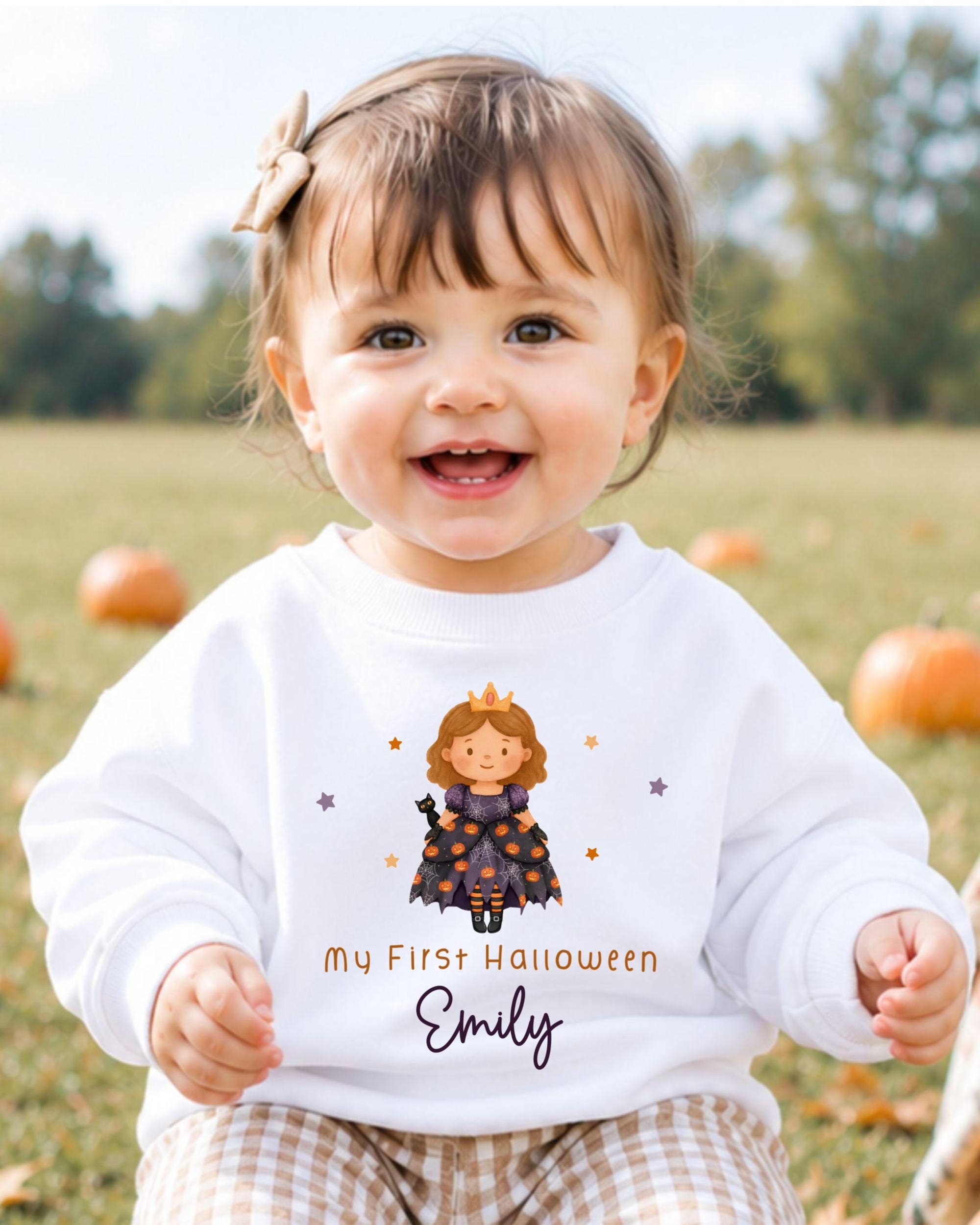 A smiling baby girl wearing a white sweater with a Halloween-themed design and text.