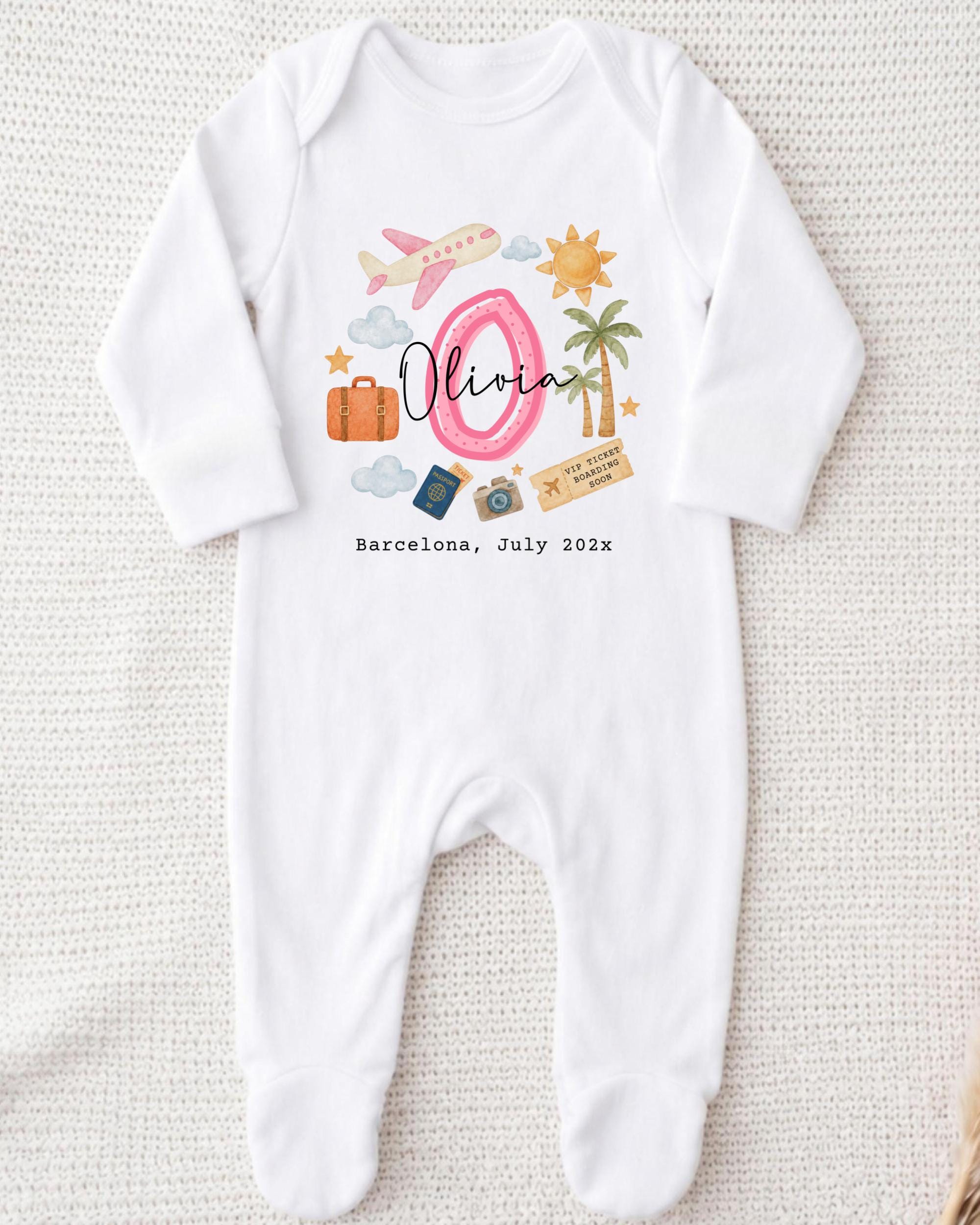A white onesie with a personalized design on it, featuring a pink number 0, a pink airplane, a palm tree, a suitcase, and a passport. The text "Olivia" is written above the design, and "Barcelona, July 2012" is written below it.