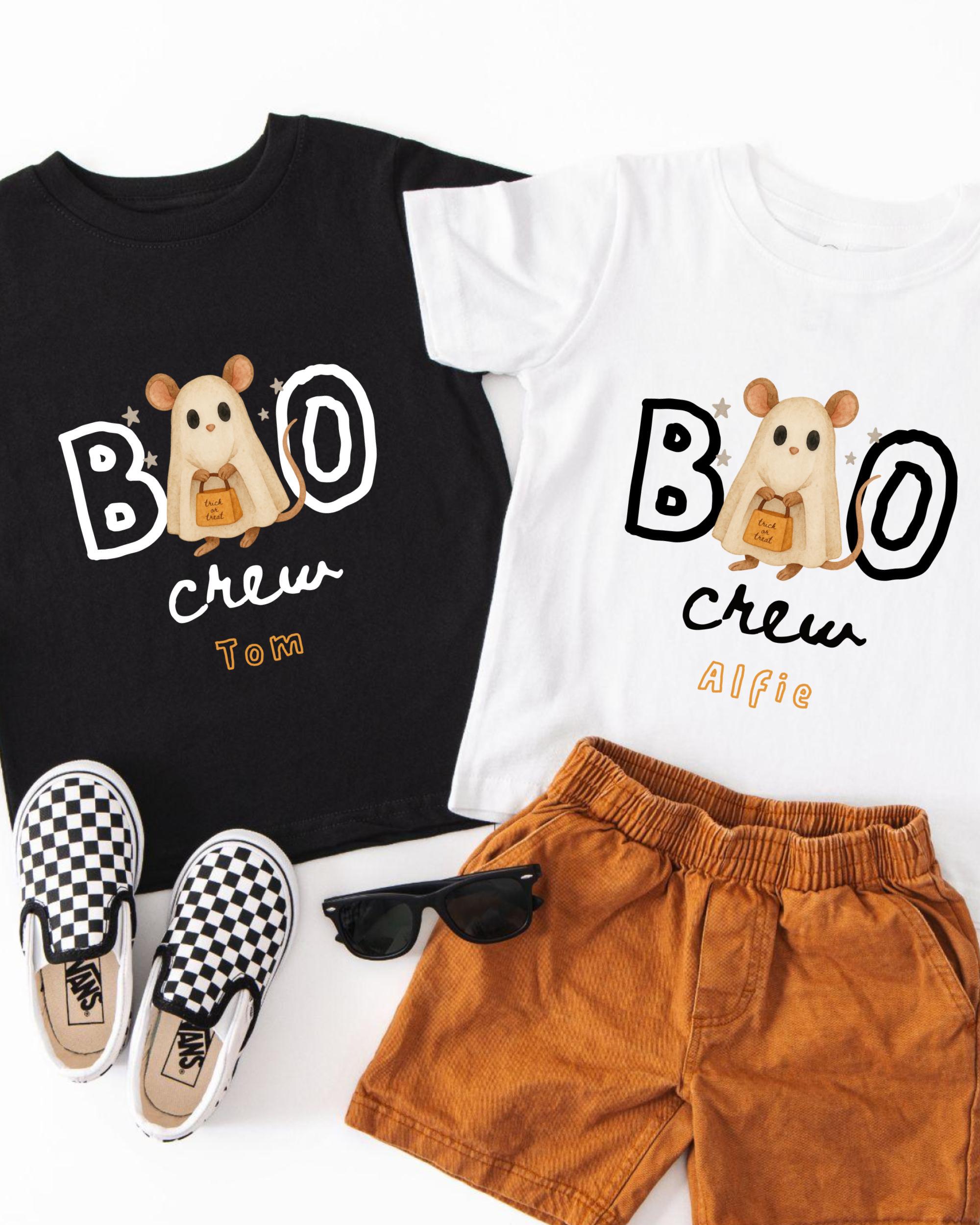A pair of children's clothing, including a black t-shirt with a ghost character and the text "BOO crew Tom Alfie", a white t-shirt with the same text, and a pair of brown shorts. There are also a pair of black and white checkered shoes and a pair of sunglasses on the floor.