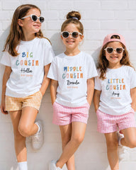 Three young girls wearing white t-shirts with personalized text and sunglasses are standing against a white brick wall.