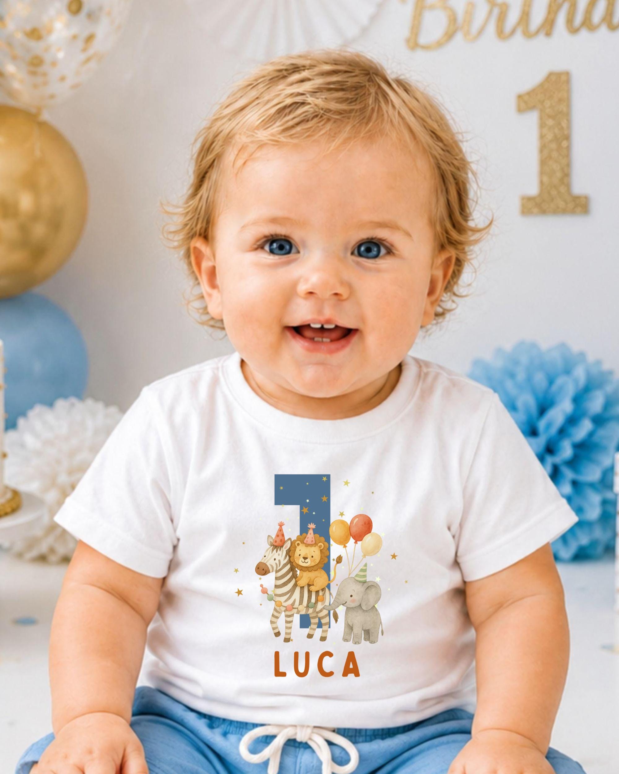 A smiling baby boy wearing a white t-shirt with a graphic of a lion, elephant, and balloons, sitting in front of a birthday party backdrop.
