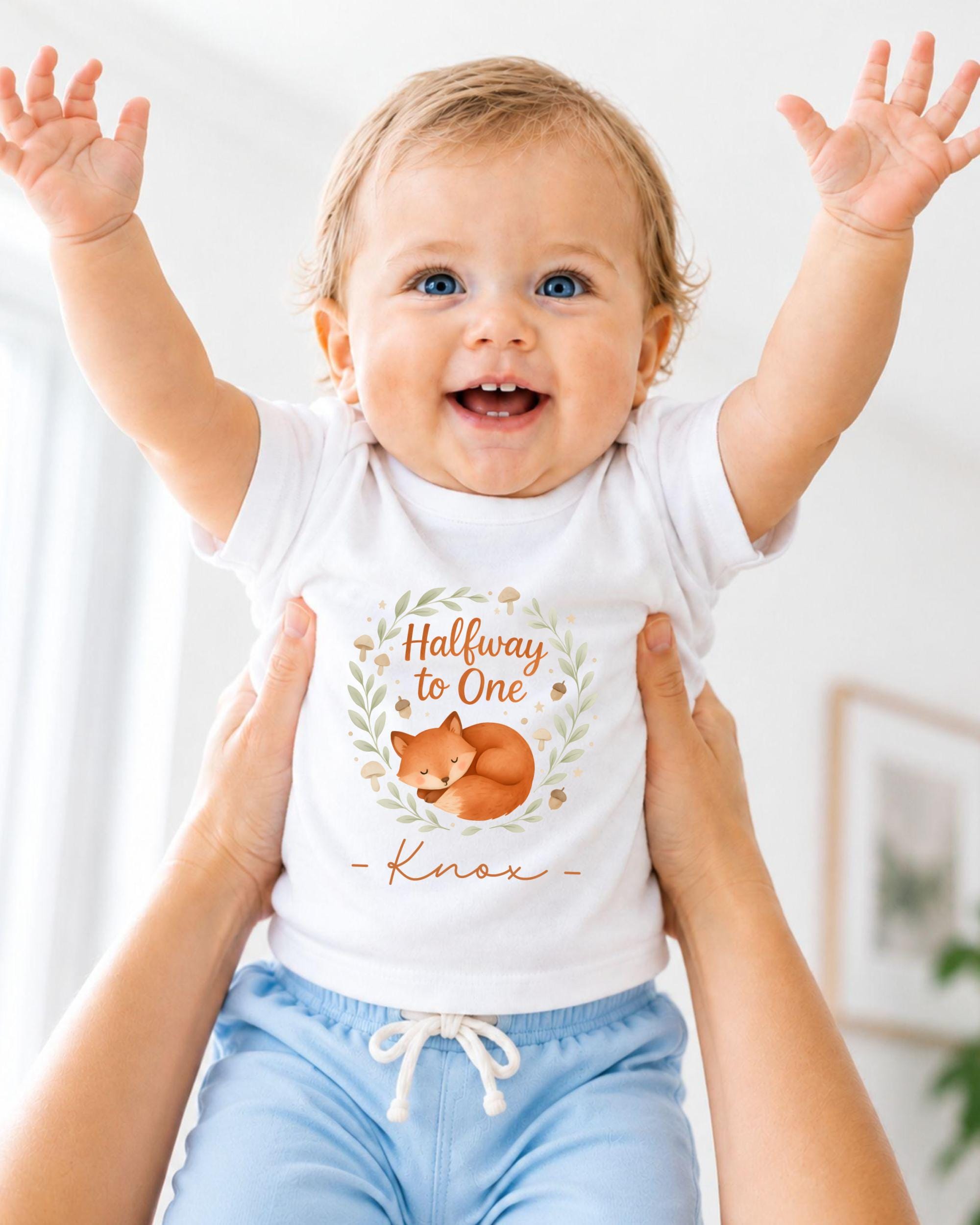 A smiling baby wearing a white t-shirt with a graphic of a sleeping fox and the text "Halfway to One" is being held up by someone's arms.