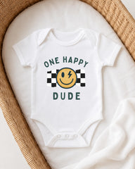 A white onesie with a smiley face and the text "ONE HAPPY DUDE" printed on it, laid out on a white blanket.