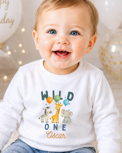 A smiling baby boy wearing a white shirt with a graphic of various animals and the text "Wild One Oscar" is the focus of the image.