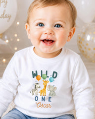 A smiling baby boy wearing a white shirt with a graphic of various animals and the text "Wild One Oscar" is the focus of the image.