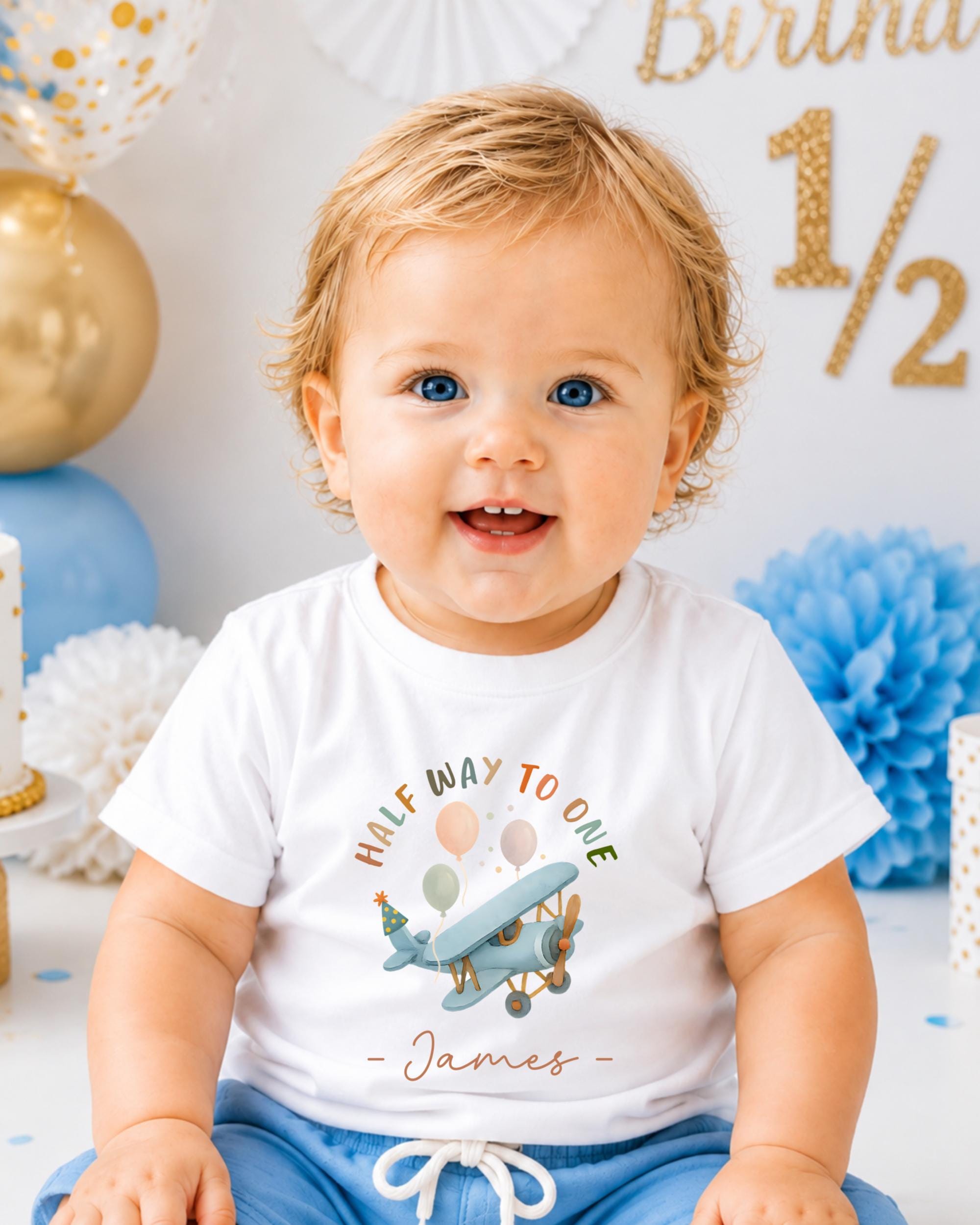 A smiling baby boy wearing a white t-shirt with a graphic of an airplane and the text "Half way to one - James" is sitting in front of a birthday party backdrop.