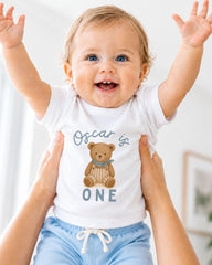 A smiling baby wearing a white t-shirt with a teddy bear graphic and the text "Oscar is one" is being held up by two people.