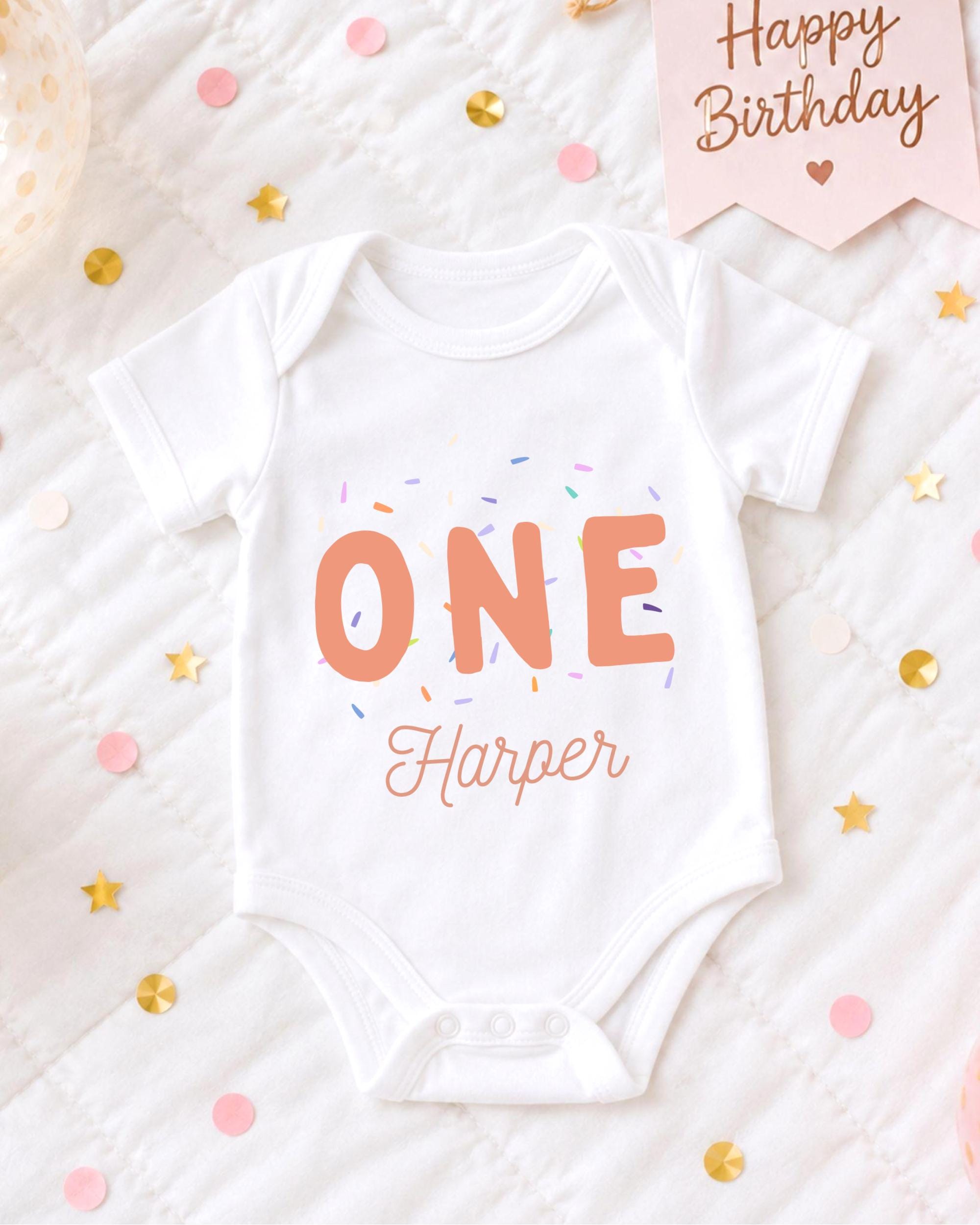 A white onesie with the text "ONE Harper" printed on it, surrounded by confetti and a birthday card.